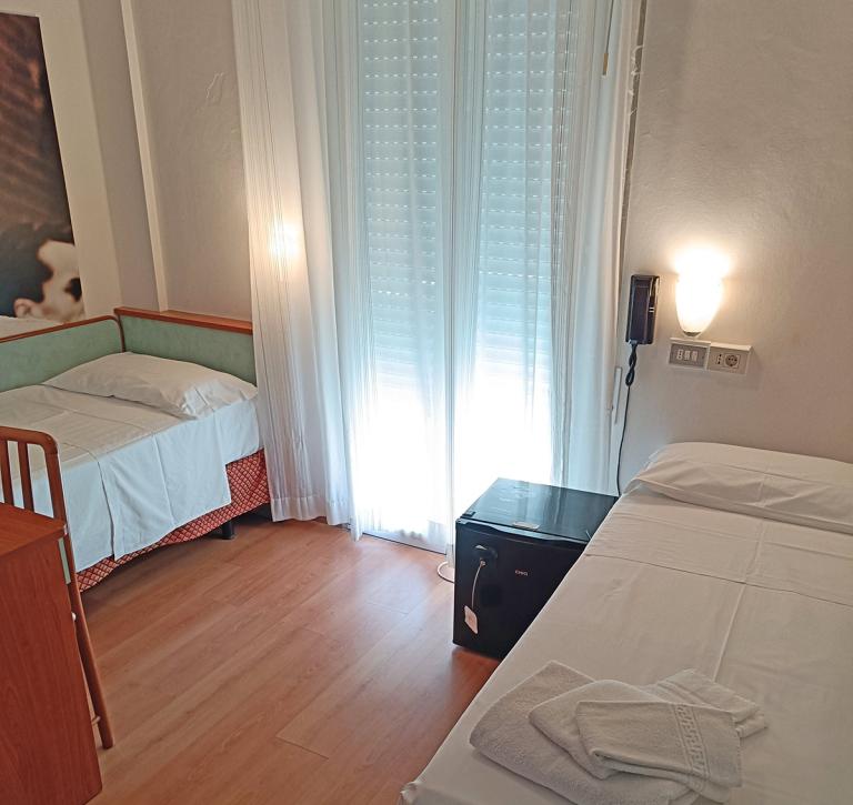 Zimmer mit zwei Einzelbetten, Wandleuchte und Fenster mit weißen Vorhängen.